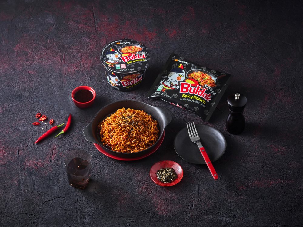 Samyang Buldak Original spicy ramen packet and cup noodle with a prepared bowl of stir-fried noodles